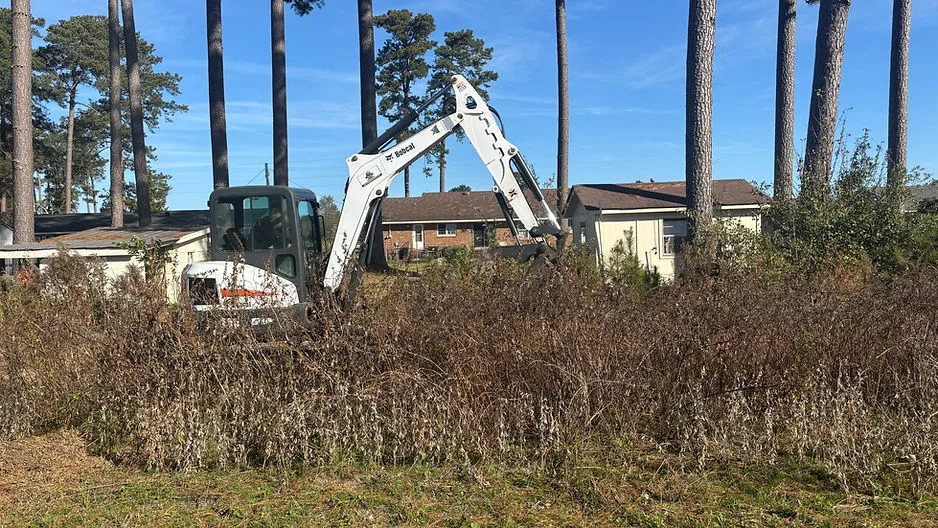 A white Bobcat excavator clearing a field of tall, brown dried weeds in front of residential houses